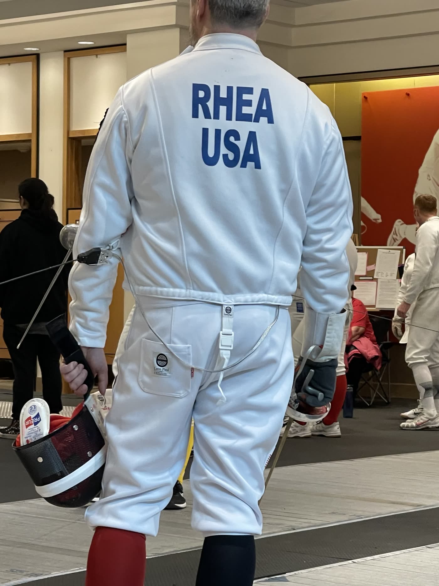 Back of Eric Rhea's fencing jacket reading RHEA USA, mask in hand between bouts